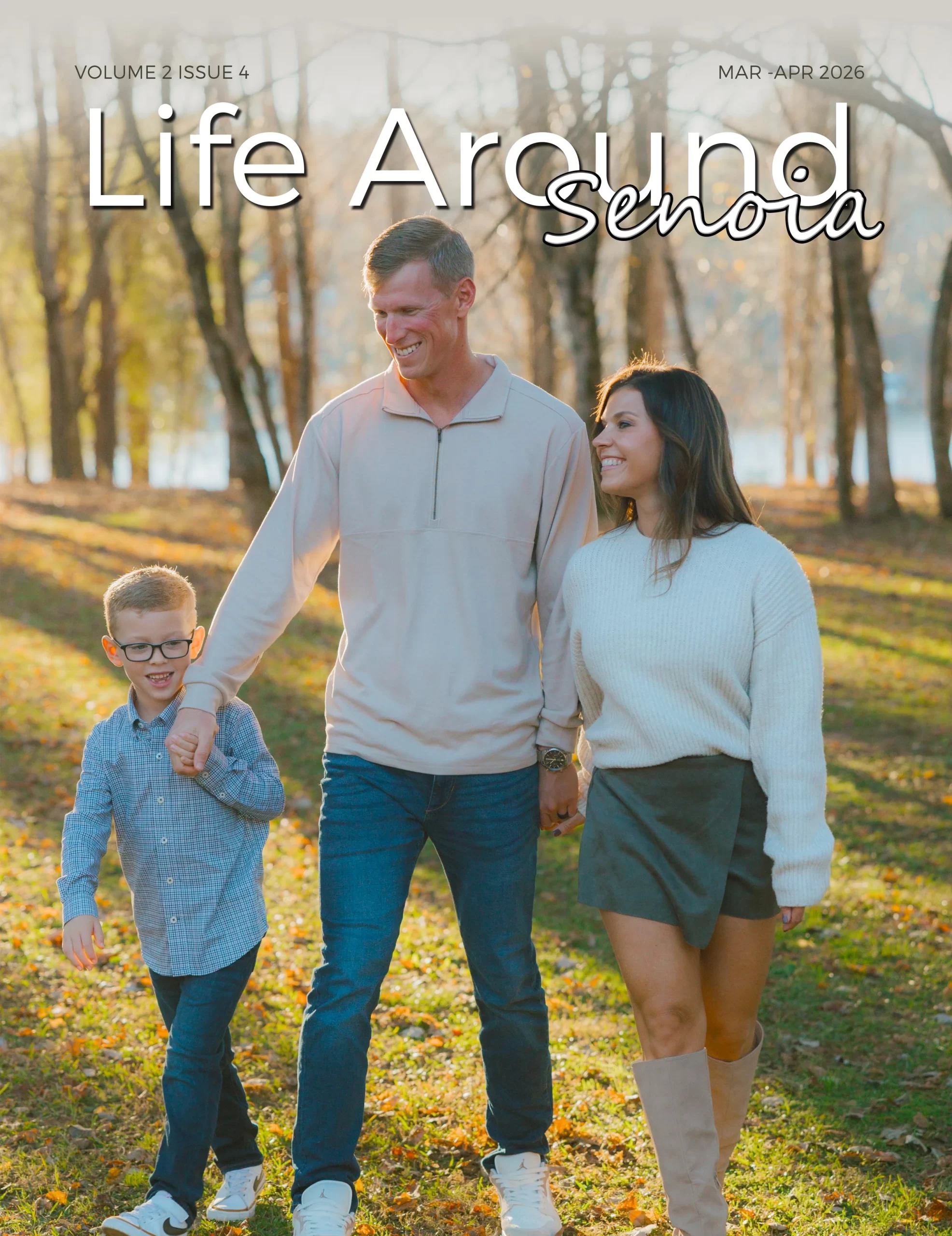 A high-resolution magazine cover for "Life Around Senoia" (Volume 2, Issue 4, Mar-Apr 2026). The photo features a smiling family of three—a father, mother, and young son—walking outdoors through a sunlit, wooded area with fallen autumn leaves on the ground. The father wears a tan quarter-zip, the mother is in a white sweater and olive skirt with tall boots, and the son wears a blue button-down and glasses. Warm, golden-hour sunlight filters through the background trees.